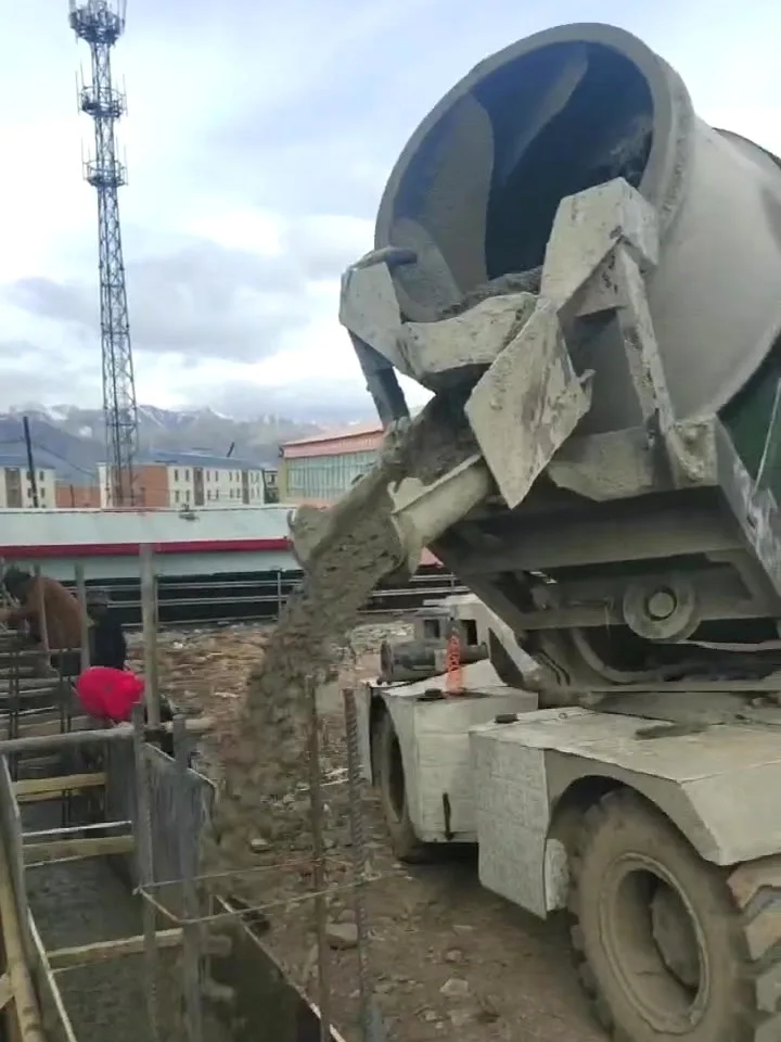 Concrete leaving a tilted mixing drum into a reinforced foundation, mountains and low-rise buildings behind