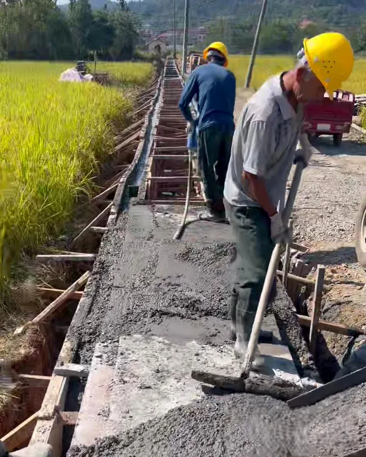 Two workers in hard hats screeding a narrow concrete strip between timber formwork beside a paddy field